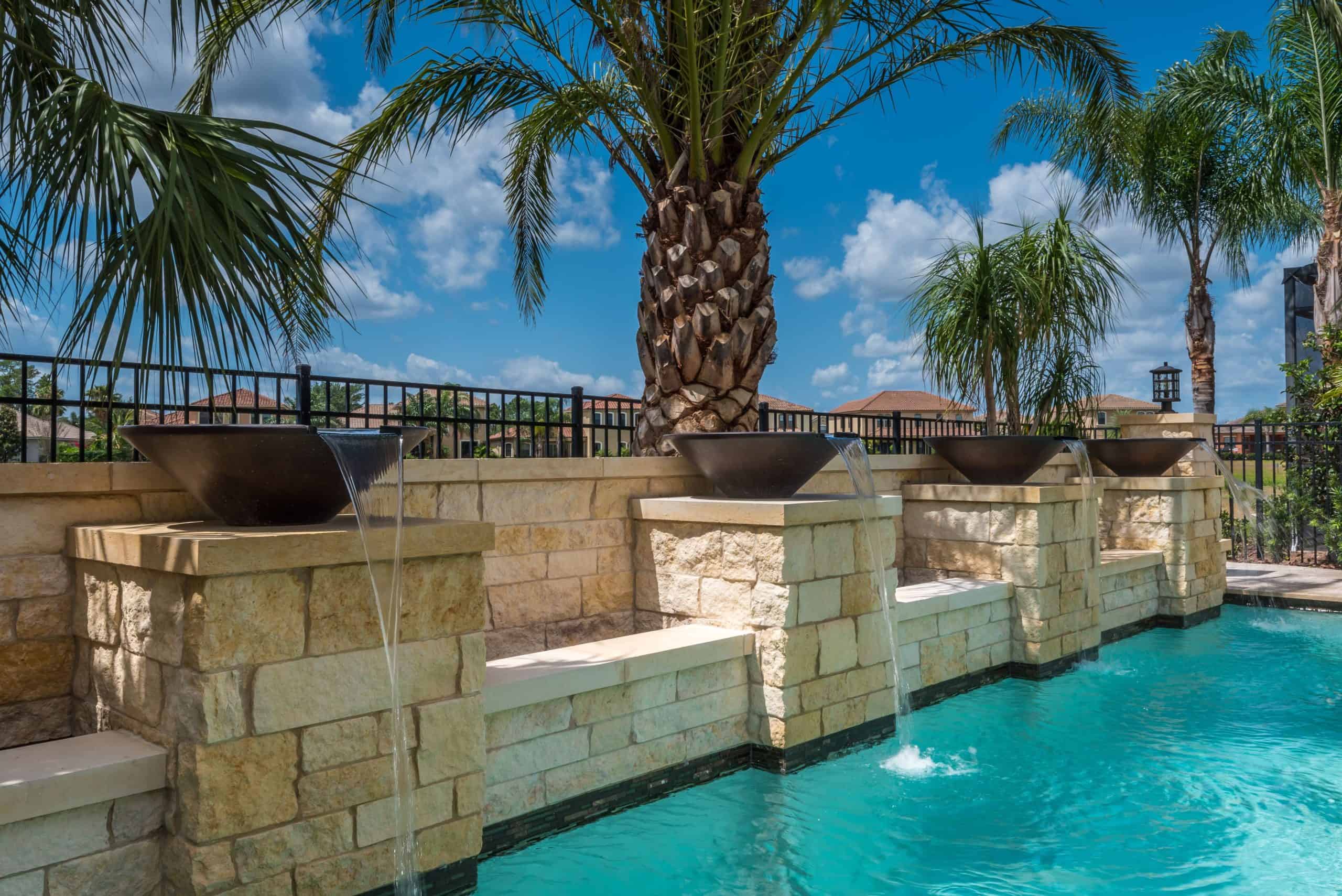 Marinus 5 of 21 Lush backyard swimming pool with stone water fountains, palm trees, and a clear blue sky.