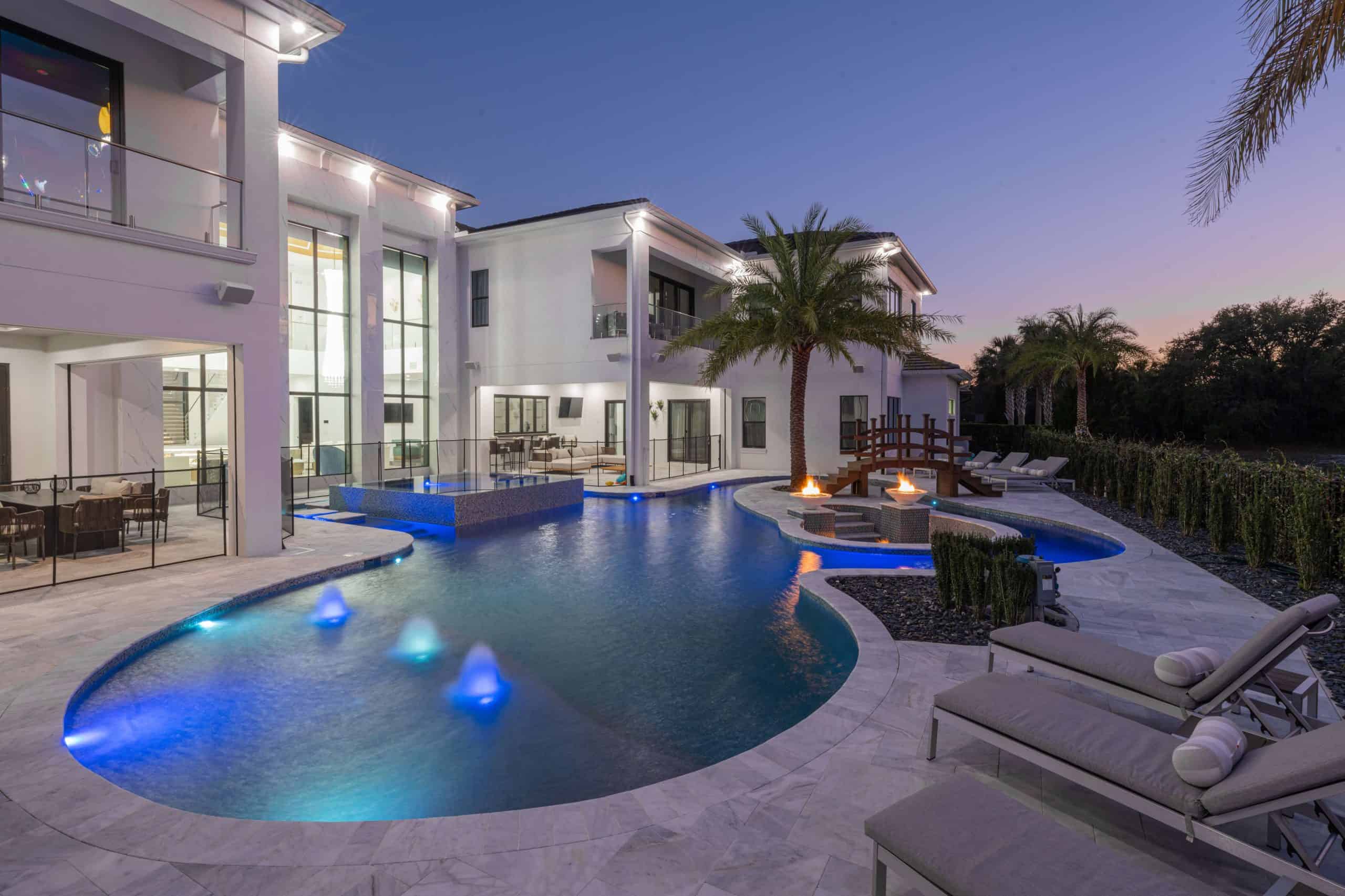 Stunning modern backyard pool with waterfall features, lounge area, fire pits, and lush palm trees at dusk in Coastal Oaks and The Outlook