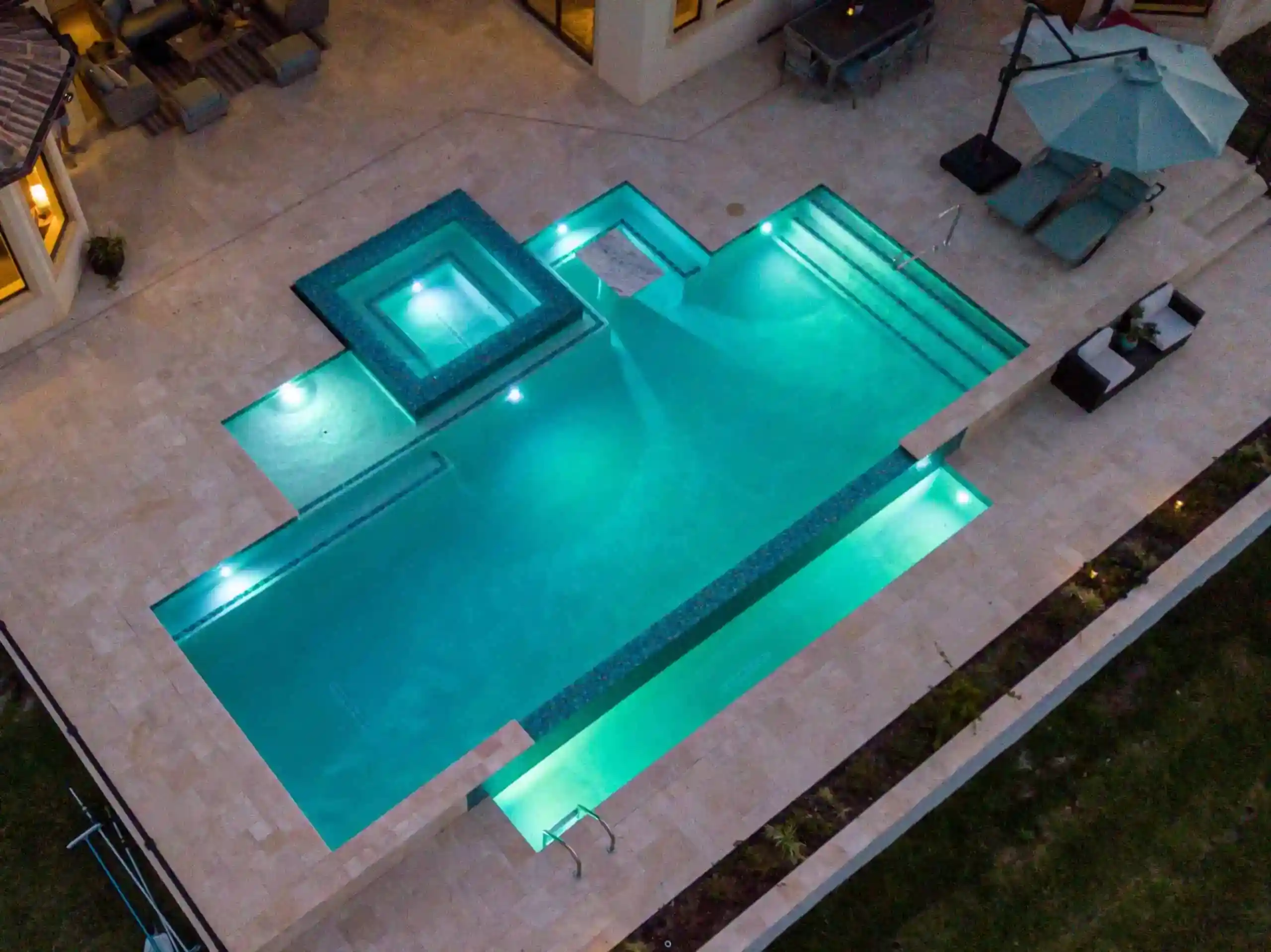 Luxurious illuminated backyard swimming pool with spa, modern design, and stylish patio area at dusk in Beachside Homes, Ormond Beach