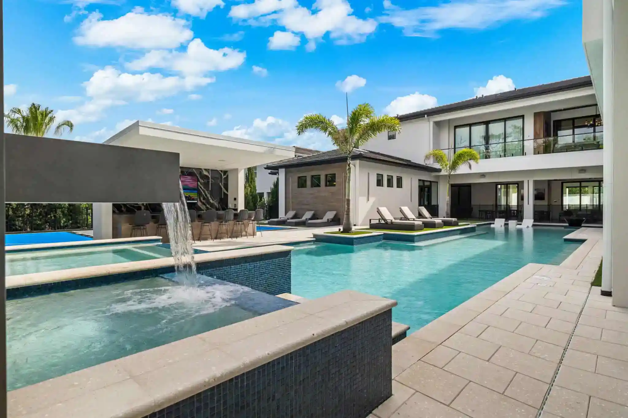 Luxurious modern backyard pool with waterfalls, lounge area, and palm trees under a blue sky in Heathrow, Seminole County FL